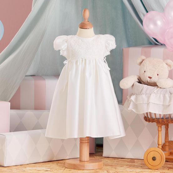 Ein weißes Kleid mit Rüschen steht auf einem Mannequin. Ein Teddybär sitzt auf einem Korb. Im Hintergrund befinden sich rosa Luftballons und ein Baldachin. Der Boden ist aus Holz.