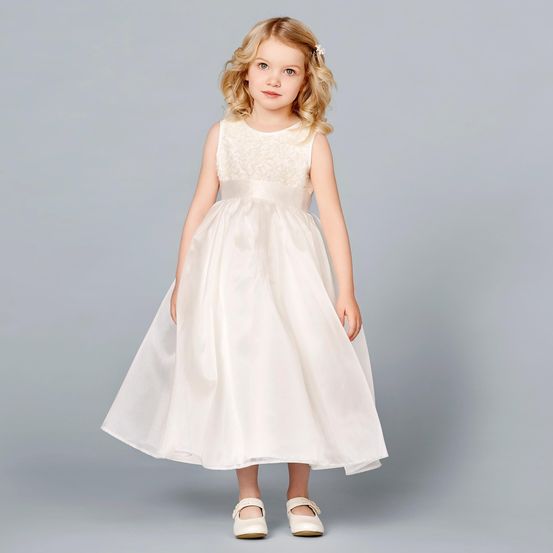 Ein kleines Mädchen mit blonden Haaren trägt ein weißes Kleid und weiße Schuhe. Sie steht vor einem grauen Hintergrund.