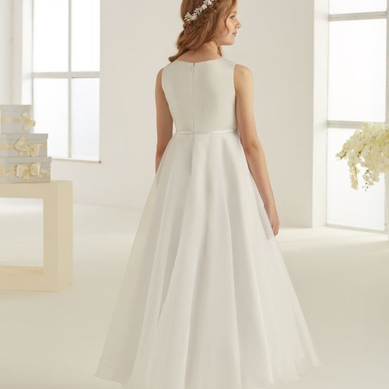 Ein Mädchen in einem weißen Kleid mit einer weißen Haarspange steht in einem Raum. Ein Tisch mit verpackten Geschenken befindet sich links. Eine weiße Blume ist rechts.