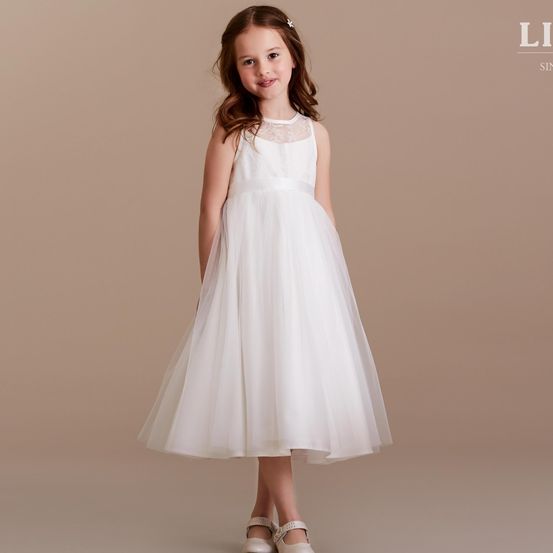 Ein kleines Mädchen in einem weißen Kleid mit Spitzendetails steht vor einem beigen Hintergrund. Sie lächelt und trägt weiße Sandalen.