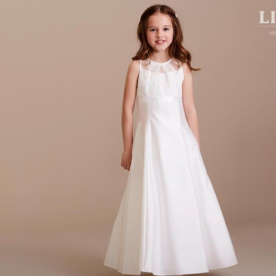 Ein junges Mädchen steht lächelnd in einem weißen Kleid mit Spitzendetails vor einem beigen Hintergrund.