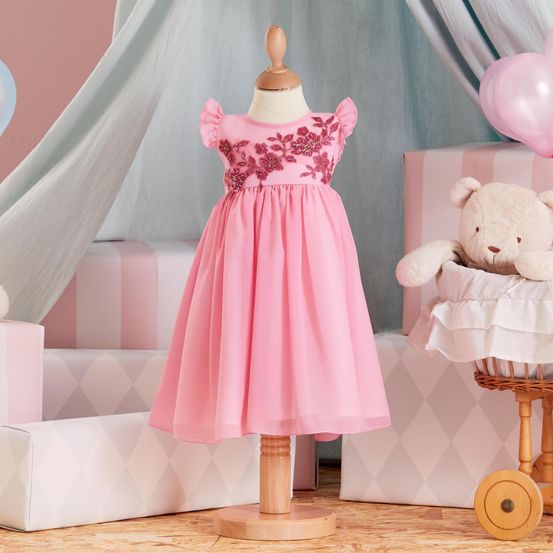 Ein rosa Kleid mit Blumenstickerei auf einem Mannequin in einem rosafarbenen Raum mit Luftballons und einem Teddybär in einem Korb.