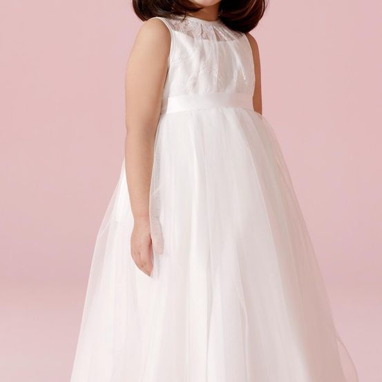 Ein junges Mädchen trägt ein weißes, ärmelloses Kleid mit Spitzendetails am Ausschnitt. Das Kleid fällt bis zum Boden mit einem vollen Rock. Sie steht vor einem rosa Hintergrund.