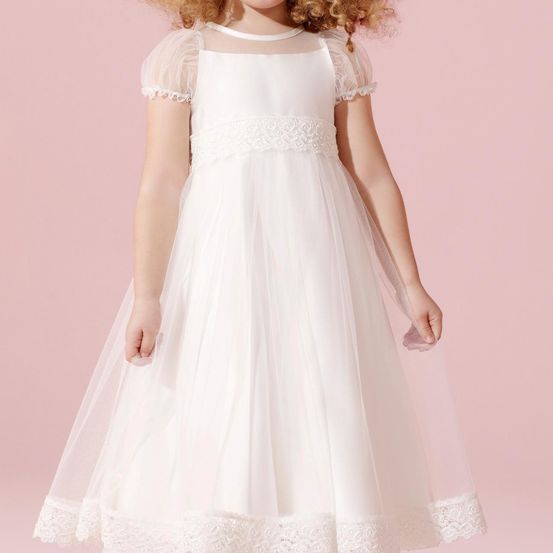 Ein junges Mädchen mit lockigen Haaren steht und trägt ein weißes Kleid mit Spitzenbesatz an den Ärmeln und dem Saum. Das Kleid hat einen durchsichtigen Oberteil und einen vollen Rock.