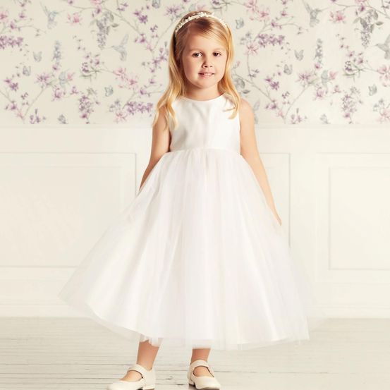 Ein kleines Mädchen in einem weißen Kleid steht in einem Raum mit Blumentapete. Sie hat eine Blumenhaarband und weiße Schuhe.