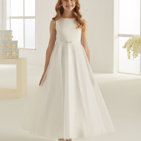 Ein junges Mädchen in einem weißen Kleid mit einer Schleife an der Taille steht in einem hellen Raum mit weißen Wänden und Fenstern.