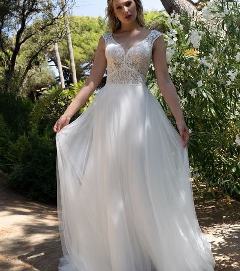 Eine Frau steht in einem Garten und trägt ein weißes Hochzeitskleid mit Spitzendetails. Der Garten ist voller verschiedener Pflanzen und Blumen.