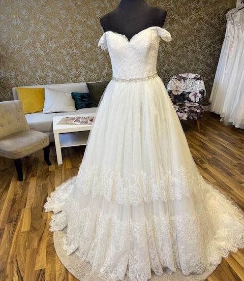 Ein weißes Hochzeitskleid auf einem Mannequin ruht auf einem runden Teppich in einem Raum mit gemusterter Tapete, einem Sofa und Stühlen.