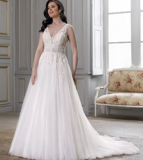 Eine Frau in einem weißen Hochzeitskleid steht in einem eleganten Raum, in der Nähe eines Fensters mit einem Blumenstuhl im Hintergrund.