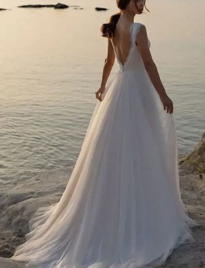 Eine Frau in einem weißen Hochzeitskleid steht bei Sonnenuntergang am Strand, mit dem Rücken zur Kamera und dem Haar zu einem Pferdeschwanz gebunden.