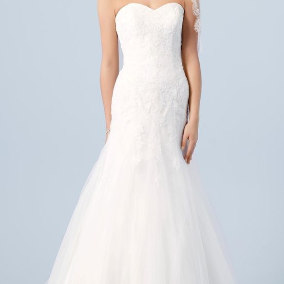 Eine Frau trägt ein ärmelloses weißes Hochzeitskleid mit Spitze und Tüll und posiert vor blauem Hintergrund.