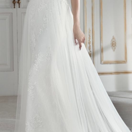 Eine Braut in einem weißen Hochzeitskleid mit Spitzendetails steht vor einer weißen Tür mit goldener Einfassung. Das Kleid hat einen vollen Rock und elegante Spitzenmuster.