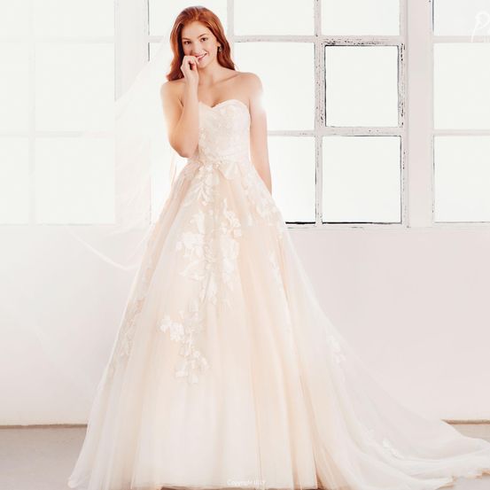 Eine Frau in einem Hochzeitskleid mit komplizierten Blumenmustern steht drinnen und lächelt in die Kamera.