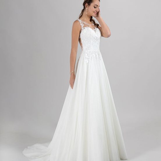 Eine Frau in einem weißen Hochzeitskleid mit aufwendigen Spitzendetails am Oberteil, die gegen einen weißen Hintergrund steht und lächelt.