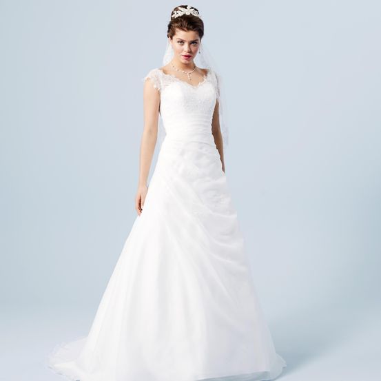 Eine Frau in einem weißen Hochzeitskleid mit Spitzendetails und einem Schleier posiert vor blauem Hintergrund. Sie trägt eine Krone und eine Halskette.