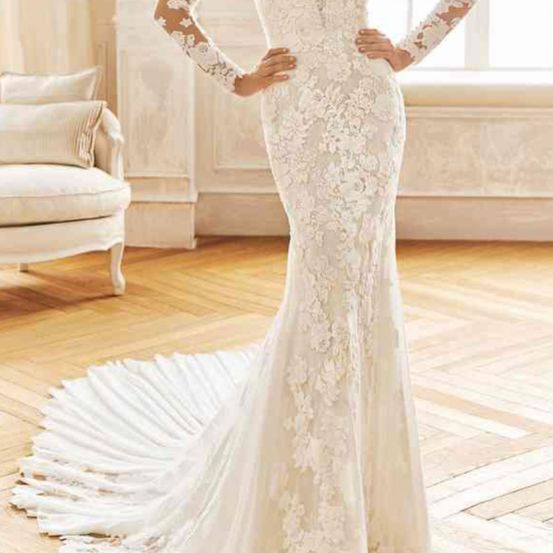 Eine Frau in einem weißen Spitzen-Meerjungfrauen-Hochzeitskleid mit langen Ärmeln steht auf einem Holzboden. Sie hat die Hände in der Taille, und ein weißer Stuhl mit Kissen steht hinter ihr.