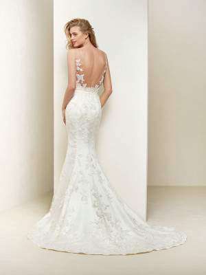 Eine Frau steht vor einer weißen Wand und trägt ein weißes Meerjungfrauen-Hochzeitskleid mit aufwendigen Spitzendesigns am Rücken. Ihr Haar ist zu einem lockeren Hochsteckfrisur gestylt.