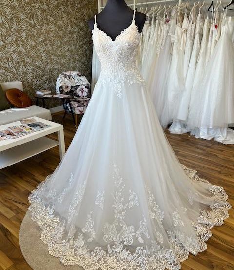 Ein weißes Hochzeitskleid mit Spitzendetails auf einem Mannequin in einem Raum mit Holzböden und einer gemusterten Wand.