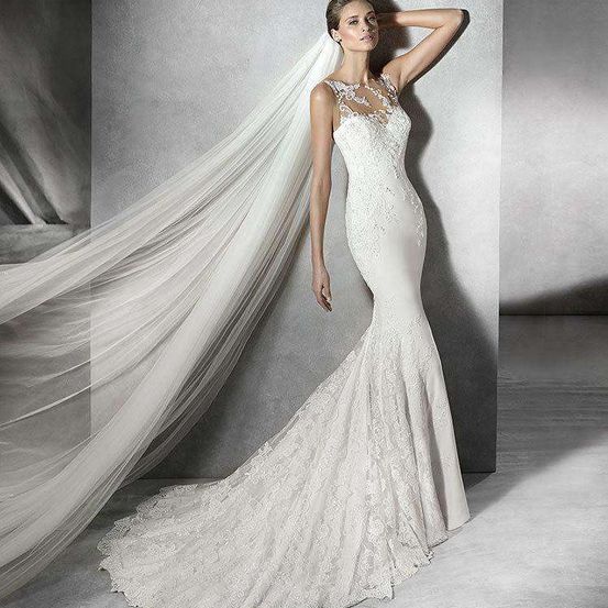 Eine Braut steht vor einer weißen Wand und trägt ein weißes Hochzeitskleid aus Spitze in Meerjungfrauenform und einen Schleier.