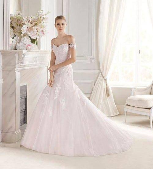 Eine Frau steht in einem Raum mit weißen Wänden und Böden, trägt ein weißes Hochzeitskleid mit aufwendigen Spitzendetails. Hinter ihr befindet sich ein Marmorkamin mit Blumen, und weiße Vorhänge bedecken ein Fenster.