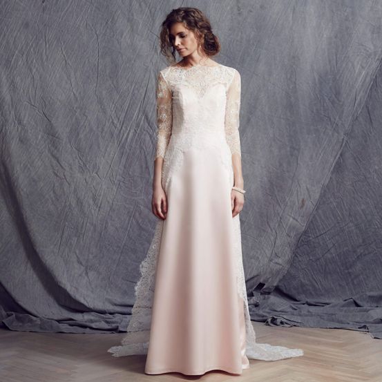 Eine Frau in einem rosa Spitzenhochzeitskleid mit langen Ärmeln und langem Schleier steht vor einem grauen Hintergrund.