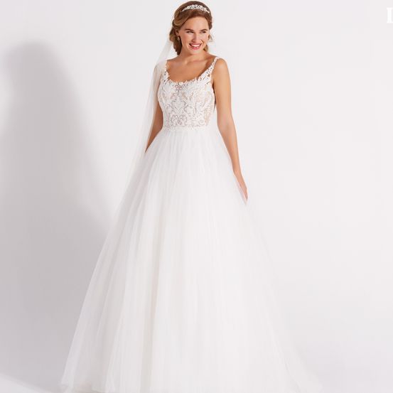 Eine Frau in einem weißen Hochzeitskleid und Schleier lächelt für ein Foto. Sie trägt eine Stirnband und steht auf einer weißen Oberfläche.