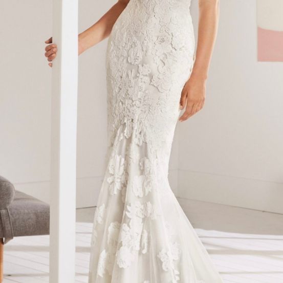 Eine Frau steht und hält einen weißen Pfeiler, trägt ein weißes Spitzenhochzeitskleid mit Meerjungfrauen-Silhouette. Das Kleid hat komplizierte Blumenmuster. Sie befindet sich in einem Raum mit weißer Wand und Boden.