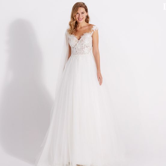 Eine Frau in einem weißen Hochzeitskleid mit Spitzendetails am Oberteil und an den Schultern posiert vor weißem Hintergrund.