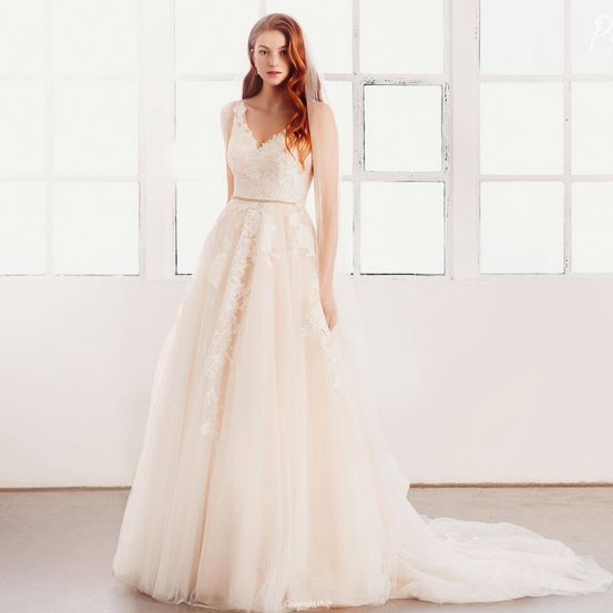 Eine Frau mit roten Haaren steht vor einem weißen Fenster und trägt ein Hochzeitskleid mit V-Ausschnitt und Spitzendetails. Das Kleid hat einen langen Schleier und sie scheint für ein Foto zu posieren.