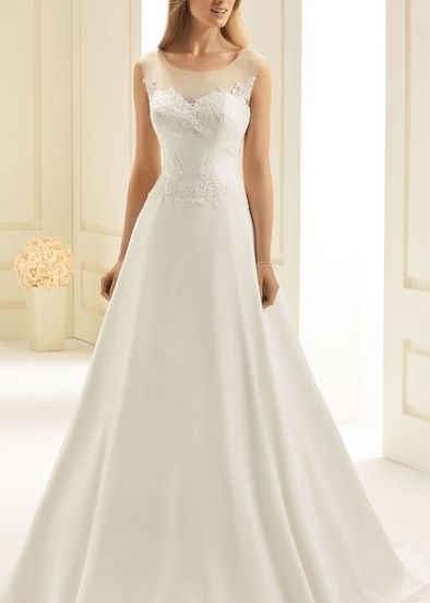 Eine Frau steht in einem Raum und trägt ein weißes Hochzeitskleid mit einer Spitzenapplikation auf dem Oberteil.