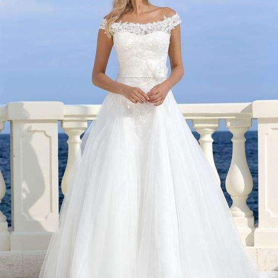 Eine Frau in einem weißen Hochzeitskleid mit offener Schulter und Spitzendetails steht auf einem Balkon mit Blick auf den Ozean.