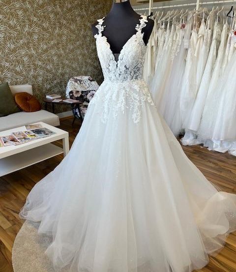 Ein weißes Hochzeitskleid mit aufwendigen Spitzendetails, das auf einem Mannequin in einem Brautgeschäft ausgestellt ist, umgeben von anderen Kleidern und Möbeln.