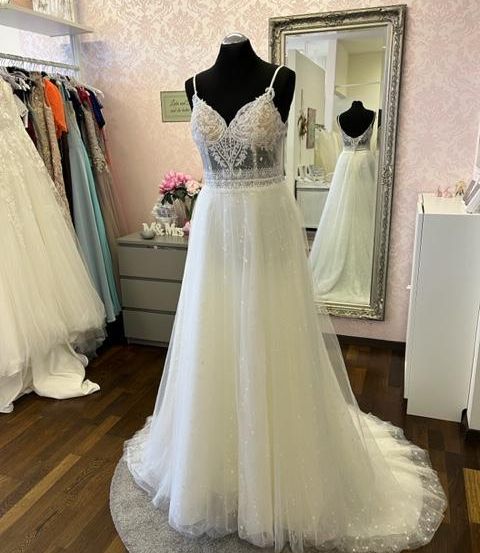 Ein Hochzeitskleid wird in einem Geschäft ausgestellt. Es steht auf einem Mannequin auf einem runden grauen Teppich. Dahinter befindet sich ein großer Spiegel, der einen Raum reflektiert.