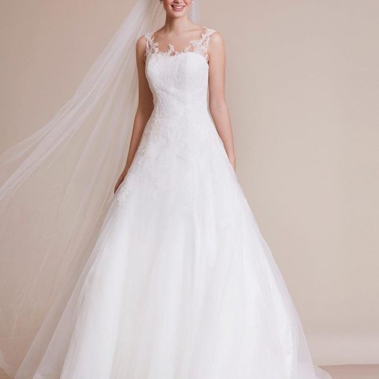 Eine Frau in einem weißen Hochzeitskleid mit Spitzendetails und einem langen Schleier steht und lächelt.