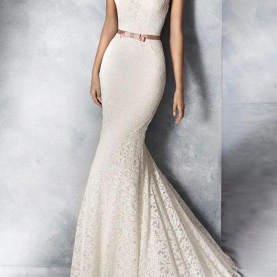 Eine Frau in einem weißen, spitzenbesetzten Meerjungfrauen-Hochzeitskleid mit einem rosa Gürtel, die an einer grauen Wand steht.