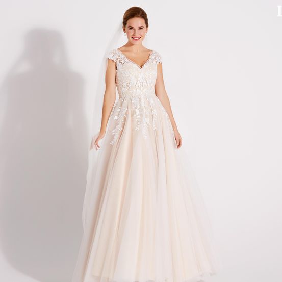 Eine lächelnde Braut in einem weißen Hochzeitskleid mit Spitzendetails und Schleier steht vor einem weißen Hintergrund.