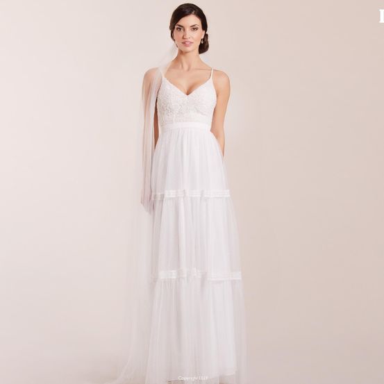 Eine Frau in einem weißen Hochzeitskleid steht mit den Händen auf den Hüften. Das Kleid hat Spitzendetails und einen langen Schleier. Der Hintergrund ist schlicht und weiß.