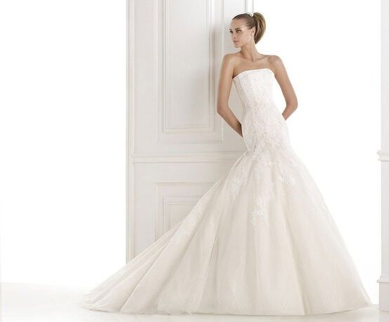 Eine Frau in einem trägerlosen weißen Hochzeitskleid mit aufwendigen Spitzendesigns steht vor einer weißen Wand.