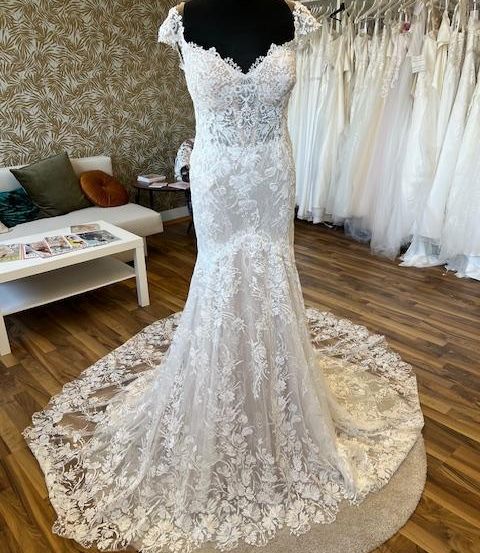 Ein Hochzeitskleid auf einem Mannequin mit aufwendigen Spitzendetails, ausgestellt in einem Raum mit Holzboden und einer Couch.