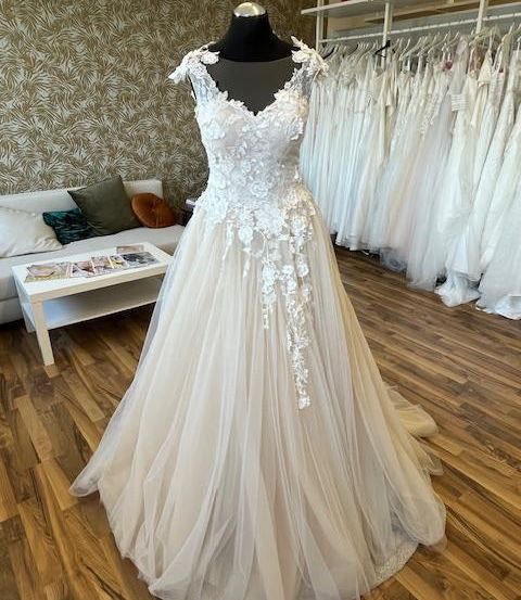 Ein Hochzeitskleid wird auf einem Mannequin in einem Brautmodengeschäft ausgestellt. Das Kleid ist weiß mit aufwendigen Spitzendetails. Der Laden hat einen Holzboden und eine weiße Couch mit Kissen.