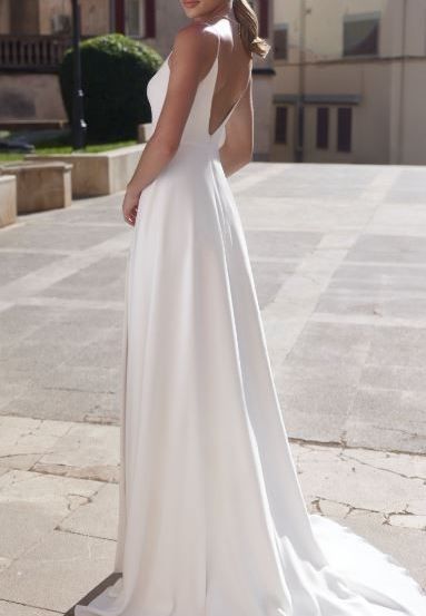 Eine Frau in einem weißen Hochzeitskleid steht auf einer Kopfsteinpflasterstraße vor einem Gebäude. Sie hat ihr Haar zu einem Dutt gebunden und sieht zur Seite.