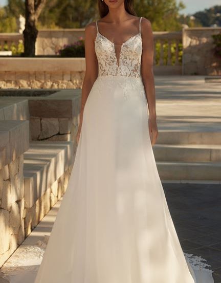 Eine Frau steht auf einem Steinweg und trägt ein weißes Hochzeitskleid aus Spitze mit tiefem V-Ausschnitt. Sie ist von Bäumen und einer Steinmauer umgeben.