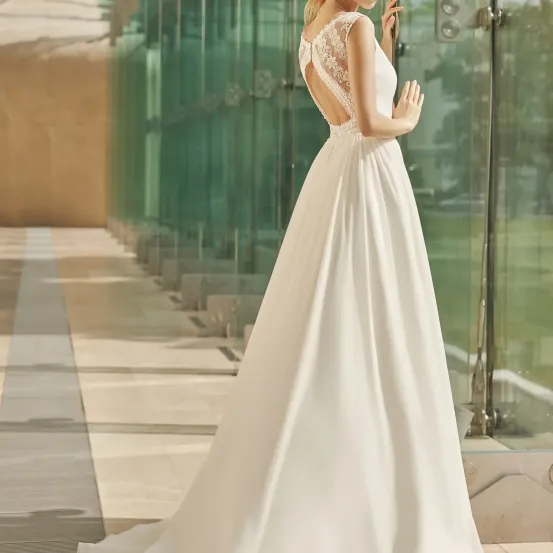 Eine Frau in einem weißen Spitzenhochzeitskleid steht vor einer Glaswand und blickt in einen Garten.