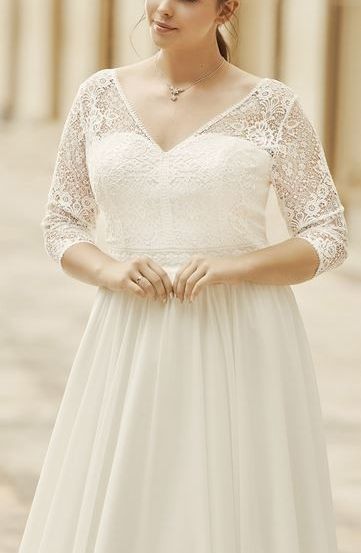 Eine Frau in einem weißen Hochzeitskleid mit Spitzenärmeln und Haarreif steht draußen, lächelt und posiert für ein Foto.