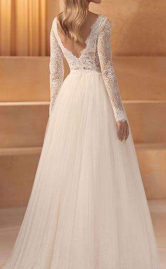 Eine Frau in einem Hochzeitskleid mit langen Ärmeln steht vor einer beigen Wand mit Stufen.