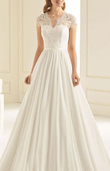 Eine Frau in einem weißen Spitzenhochzeitskleid mit V-Ausschnitt und Ärmelchen posiert drinnen. Sie hat eine Blume im Haar und eine Halskette an.