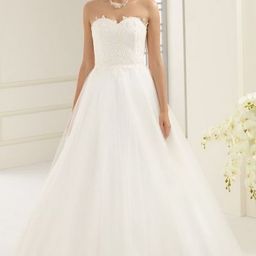 Eine Frau in einem weißen Hochzeitskleid steht in einem Raum mit weißen Wänden und einem Fenster. Sie trägt eine Halskette und hat blonde Haare. Sie schaut nach vorne.