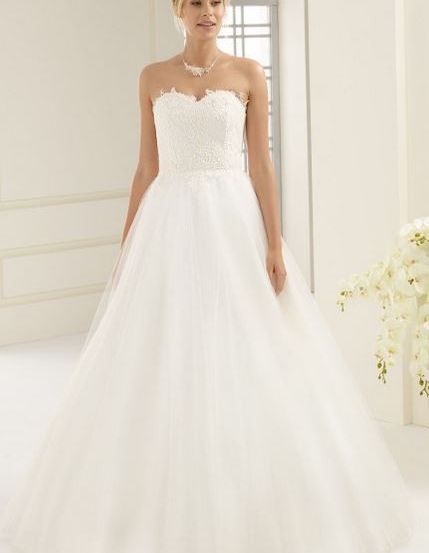 Eine Frau in einem weißen Hochzeitskleid steht in einem Raum mit weißen Wänden und einem Fenster. Sie trägt eine Halskette und hat blonde Haare. Sie schaut nach vorne.