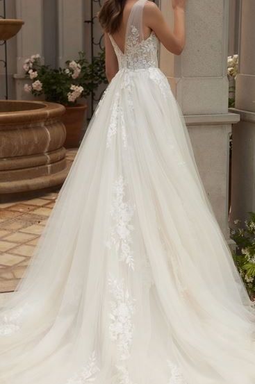 Eine Frau posiert in einem weißen Hochzeitskleid mit kunstvollen Spitzen und einem langen Schleier, neben einer Säule mit dekorativen Pflanzen im Hintergrund.