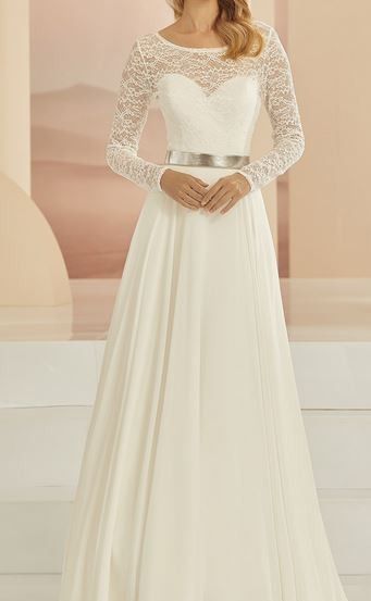 Eine Frau in einem weißen Hochzeitskleid mit langen Ärmeln und Spitzendetails, die auf einer Bühne steht, wahrscheinlich bei einer Hochzeit.